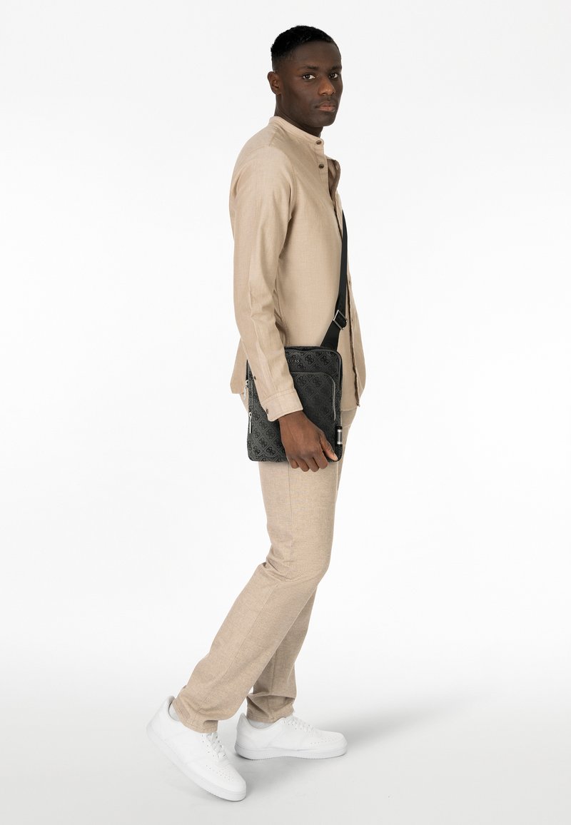 Borsa messenger nera con texture e design a motivo, indossata a tracolla, abbinata a una camicia di lino beige e pantaloni coordinati. Scarpe da ginnastica bianche.