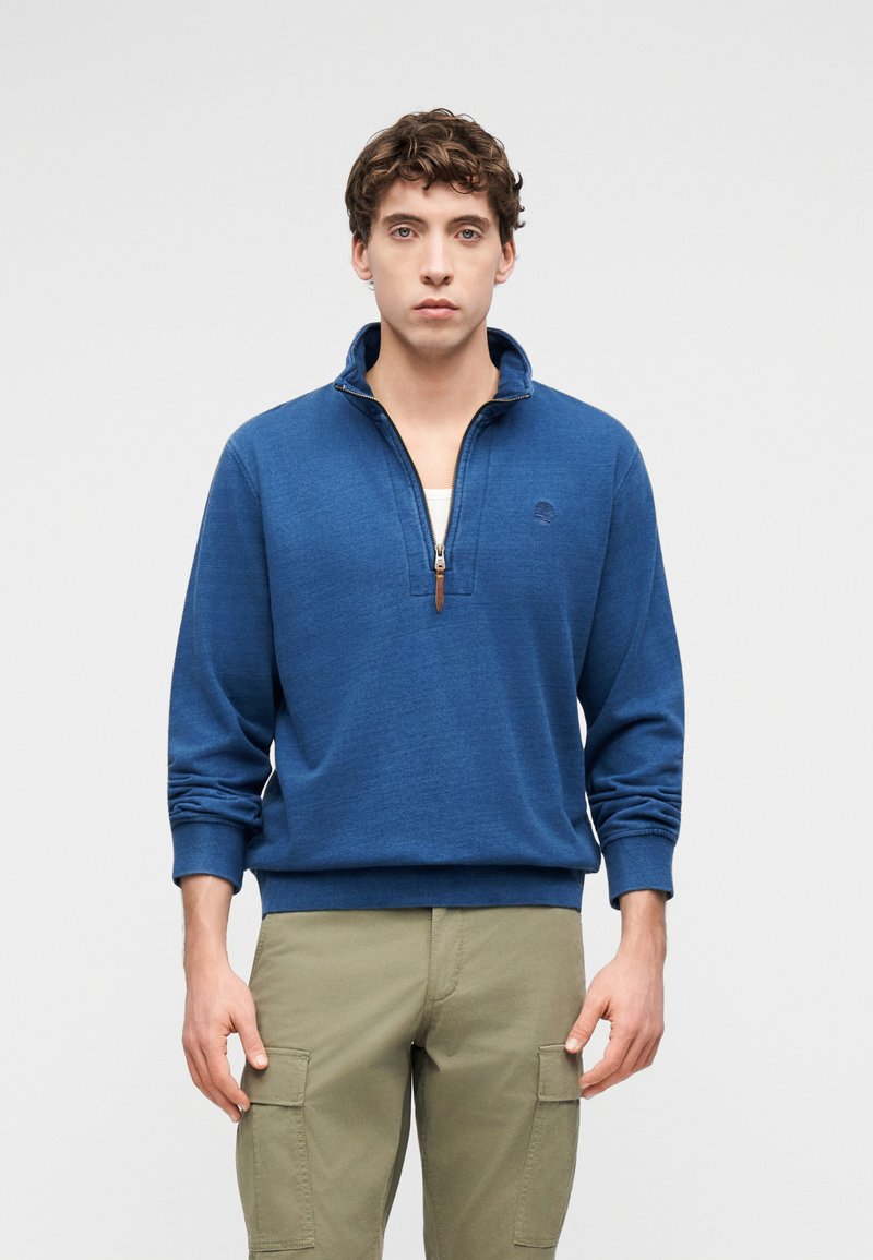 Jeune homme portant un pull bleu à demi-fermeture éclair par-dessus une chemise blanche et un pantalon cargo beige, debout devant un fond uni.