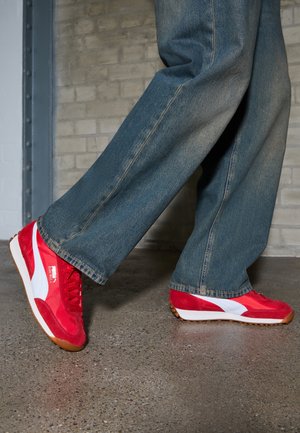 Personne portant un jean bleu et des baskets Puma rouges et blanches marchant sur un sol en béton poli devant un mur de briques.
