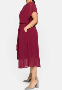 Robe midi bordeaux à manches courtes, avec poches latérales et taille nouée. Fabriquée en tissu léger et transparent ; associée à des chaussures à talons carrés grises.