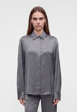 Jeune femme aux cheveux lisses portant une chemise en satin grise à manches longues boutonnée et un pantalon assorti sur un fond uni.