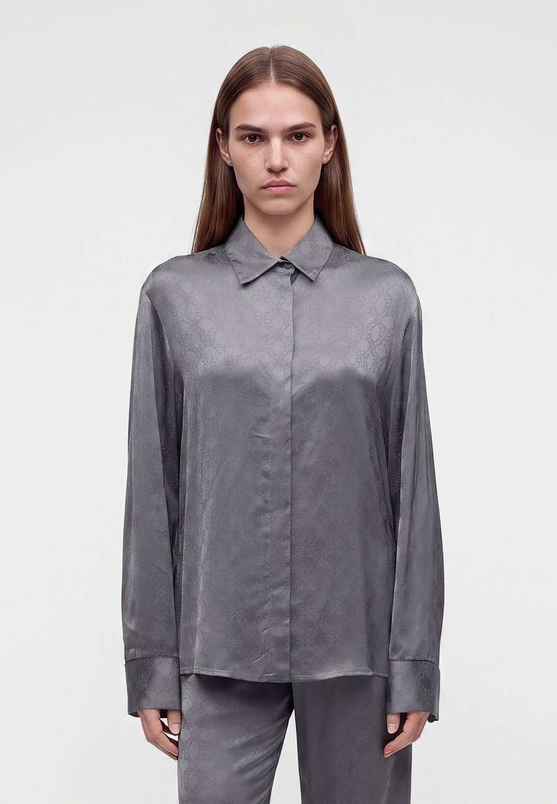 Jeune femme aux cheveux lisses portant une chemise en satin grise à manches longues boutonnée et un pantalon assorti sur un fond uni.