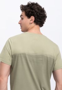 Olive green t-shirt with short sleeves, featuring a gradient design on the back and a subtle scissor logo at the neckline.