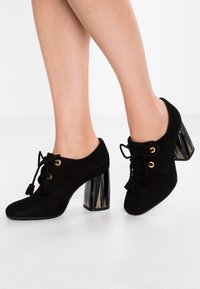 Svarta mockaskor med rund tå, snörning fram, gyllene öglor och texturerade klackar i blockstil med ett mönster i svart och klart.