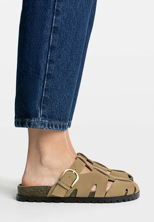 Stopy w beżowych zamszowych sandałach zakrytych, z klamrami i czarnymi podeszwami, połączone z krojonymi ciemnoniebieskimi dżinsami.