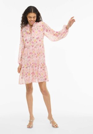 Fiatal nő mosolyogva, rózsaszín virágos hosszú ujjú ruhát és ezüst tűsarkú szandált viselve, egyik karját felemelve pózol fehér háttér előtt.