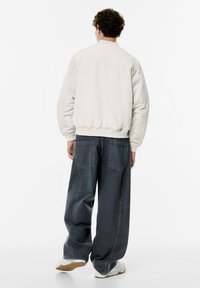 Light beige bomber jacket with ribbed hem and cuffs, paired with loose-fit dark denim pants featuring two back pockets. White sneakers.