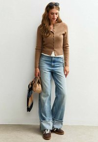Une femme se tient à côté d'un mur blanc, portant un cardigan marron, une chemise blanche, un jean large et des chaussures marron, tenant un petit sac et une écharpe.