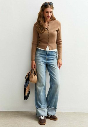 En kvinna står bredvid en vit vägg, klädd i en brun kofta, en vit skjorta, breda jeans och bruna skor, medan hon håller en liten väska och en scarf.