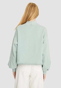 Sudadera verde claro con un cuello alto y mangas abullonadas, hecha de un material suave, de longitud cropped y con detalles de costura mínimos.