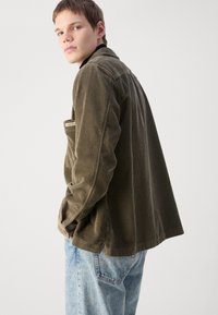 Dark olive corduroy jacket with a relaxed fit, featuring a pocket and subtle branding on the left side, paired with light blue jeans.