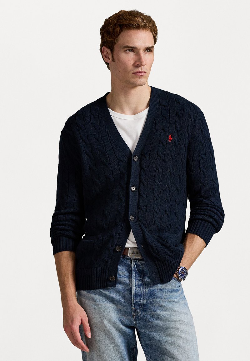 Ναυτικό μπλε καφετί ζακέτα με πλέξιμο, με κουμπωμένο μπρ�οστινό μέρος, ριμπ μανσέτες και χαμηλή ραφή, με μικρό κόκκινο λογότυπο στο αριστερό στήθος. Φορεμένο πάνω από λευκό πουκάμισο.