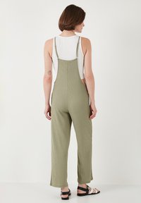 Tutina in cotone verde oliva con gamba ampia, spalline regolabili e tessuto testurizzato. Indossata sopra una canotta bianca, abbinata a sandali neri.