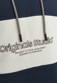Sudadera a rayas en azul marino y crema, con tejido texturizado, cordones con acentos en negro y un logo que dice "Originals Studio" en un lugar destacado.
