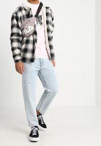 Person iført lyseblå jeans, jakke med sort-hvidt tern, pink grafisk t-shirt, sorte sneakers og grå FILA crossover taske.