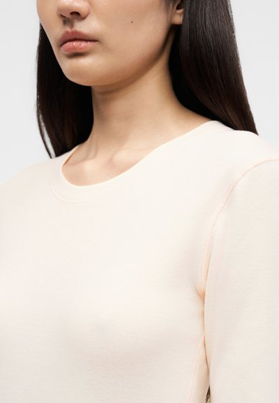 Νεαρό άτομο με μακριά σκούρα μαλλιά, που φοράει μια ανοιχτόχρωμη ροδακινί μπλούζα με μακριά μανίκια και στρογγυλή λαιμόκοψη, εμφανιζόμενο από τους ώμους μέχρι το πηγούνι.