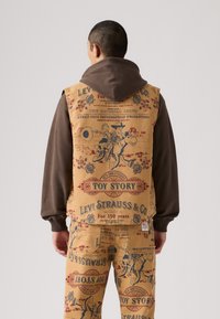 Bež prsluk i odgovarajuće hlače sadrže otisnute grafike iz vintage "Toy Story", s smešnim naglascima i džepovima. Materijal deluje čvrsto i mekano.