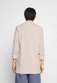 Blazer beige sur mesure avec manches trois-quarts, une fente au dos et une texture lisse, associé à un pantalon bleu ample.