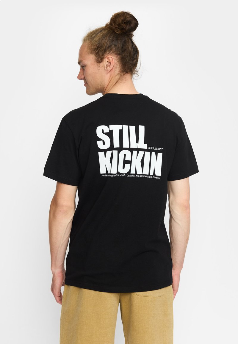 Un homme avec un chignon de cheveux bruns porte un t-shirt noir avec le texte blanc en gras "STILL KICKIN" dans le dos et un pantalon beige, se tenant devant un fond blanc.