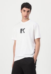 T-shirt de algodão branca com um logo gráfico preto na frente. Design de mangas curtas e decote redondo. Textura suave e ajuste relaxado.