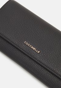 Portafoglio in pelle nera con superficie texture, caratterizzato da un logo "Coccinelle" in tono oro. Design slim e linee pulite con chiusura a pattina.