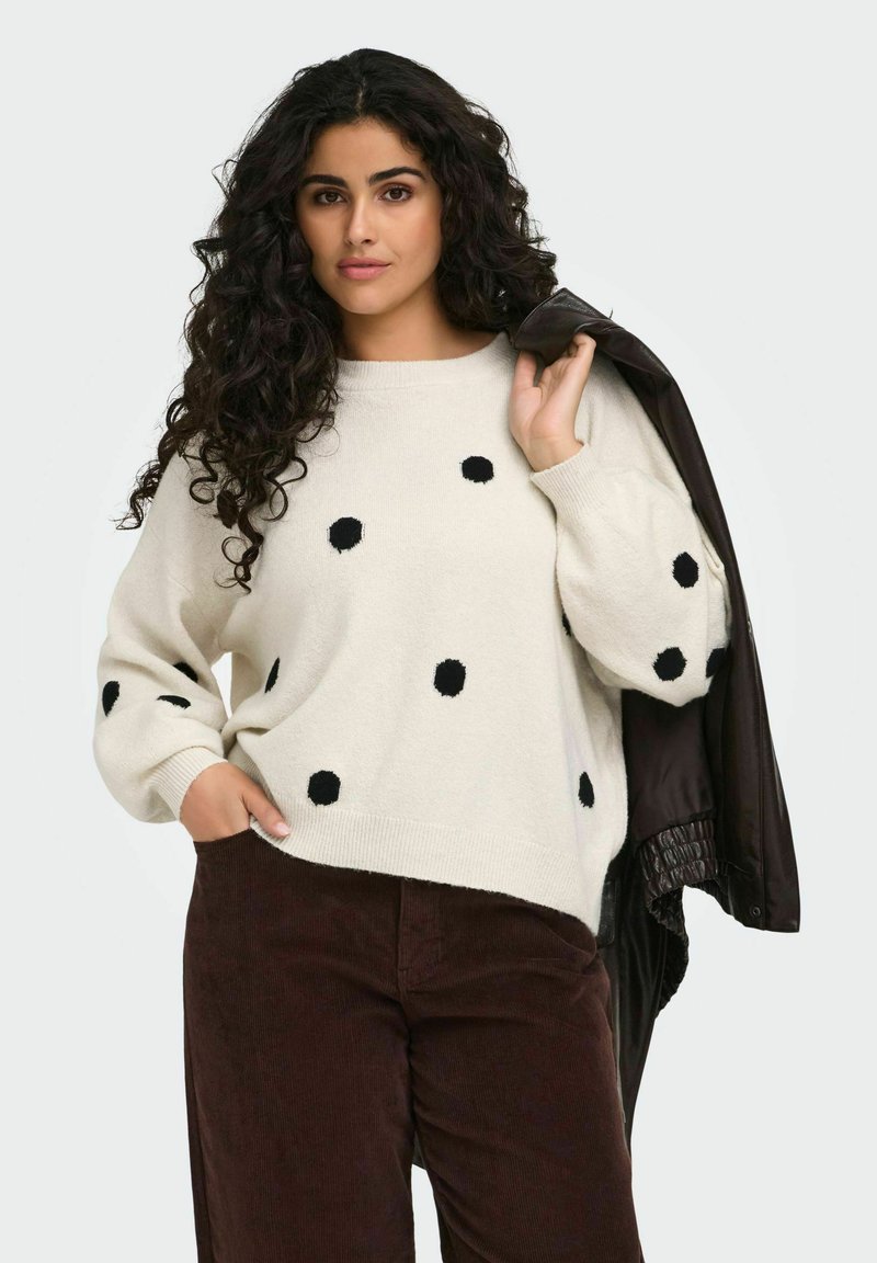 Femme aux cheveux bouclés et foncés porte un pull blanc à pois noirs, un pantalon marron, et tient une veste noire sur l'épaule.