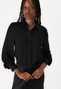 Black, long-sleeve blouse with vertical stripes, button-up front, and relaxed cuffs, paired with a smooth black waistband skirt.