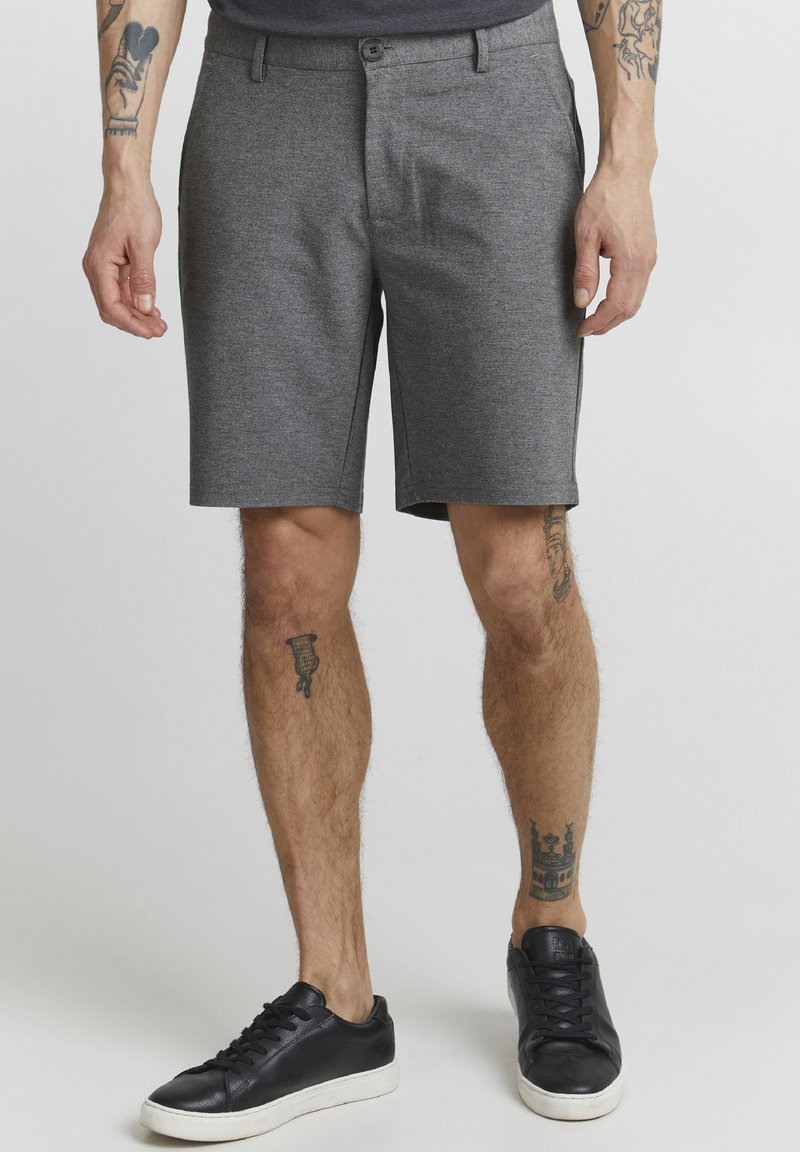 Shorts taillés gris en tissu texturé, avec une fermeture à bouton sur le devant et des poches latérales, associés à des baskets noires.