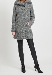 Donna che indossa un cappotto grigio con texture e cappuccio, leggings in pelle nera e stivaletti neri con tacco alto, in piedi contro uno sfondo semplice.