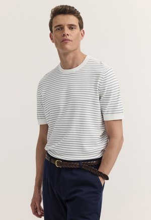 Mann, der ein weißes Kurzarmhemd mit dünnen schwarzen Streifen, dunkelblaue Hose und einen geflochtenen braunen Gürtel trägt, steht mit einer Hand in der Tasche.