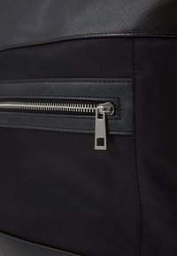 Borsa di stoffa nera con cerniera argento, dotata di un pannello superiore texture e una sezione inferiore liscia. Il tirante della cerniera presenta un dettaglio a taglio quadrato.