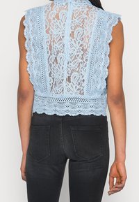 Ljusblå spetsblus med vågiga kanter och blommönster. Bakstycket har ett detaljerat genomskinligt spets. Kombinerad med mörka jeans.