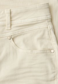 Primer plano del bolsillo de jeans de mezclilla beige con presilla para cinturón, doble costura y remaches metálicos sobre la textura de la tela.