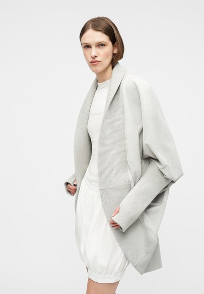 Γυναίκα που φοράει υπερμέγεθη ανοιχτό γκρι ζακέτα με φαρδύ γιακά πάνω από λευκό φόρεμα, ποζάροντας μπροστά σε απλό ανοιχτό φόντο.