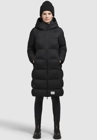 Zwart gewatteerde lange jas met een ritssluiting, capuchon en zijzakken. Draag het over zwarte leggings en enkellaarsjes. Heeft een logo-opdruk aan de onderrand.