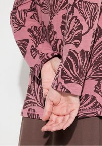 Kädet ristissä selän takana, päällä vaaleanpunainen pitkähiha top, jossa on tummia kukkakuoseja ja ruskeat housut, yksinkertaista taustaa vasten.