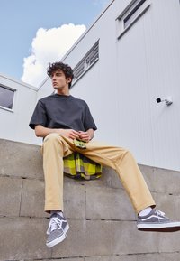 T-shirt gris, pantalon beige, baskets gris et blanches. Tenant un objet vert, assis sur un mur en pierre. Chemise à carreaux drapée derrière.