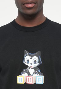 Black cotton t-shirt featuring a cartoon cat design with blue and white accents, sitting above colorful building blocks spelling "HUF."