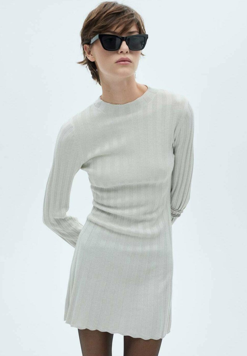 Hellgraues, geripptes Kleid mit langen Ärmeln und gewelltem Saum. Das Model trägt übergroße schwarze Sonnenbrillen, die einen kontrastierenden Akzent setzen.