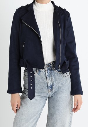 Kvinde iført marineblå kort jakke med lynlås og bælte over en hvid ribbet rullekrave, kombineret med lyseblå højttaljede jeans.