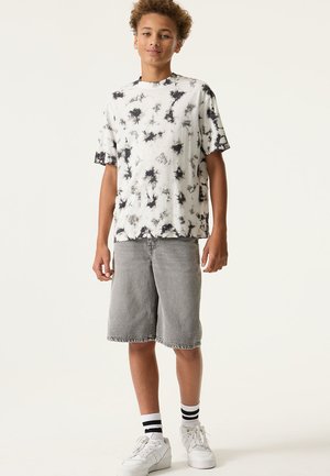 Weißes und schwarzes Batik-T-Shirt mit kurzen Ärmeln, kombiniert mit grauen Denim-Shorts, weißen Turnschuhen und schwarz-weiß gestreiften Socken.