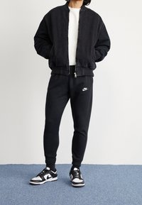 Černá bomber bunda, bílé tričko s kulatým výstřihem, černé tepláky s logem Nike a černobílé tenisky. Ležérní, přiléhavý střih, hladká textura.