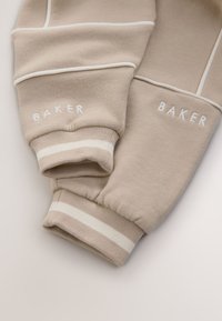 Poignets de pantalon de jogging beige avec des rayures blanches et un détail de passepoil blanc, avec le mot ""BAKER"" brodé en blanc sur chaque poignet.