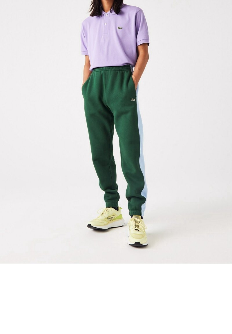 Gröna sweatpants med blå sidopaneler, tillsammans med en lavendelfärgad polo-tshirt. Har en logotyp på både t-shirten och sweatpantsen. Gula sneakers.