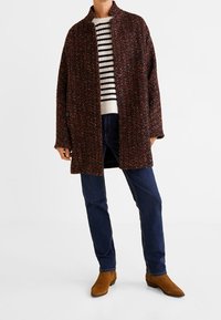 Abrigo marrón texturizado con un collar, cierre de botones, usado sobre un suéter de rayas. Combinado con jeans oscuros y botas de tobillo marrones.