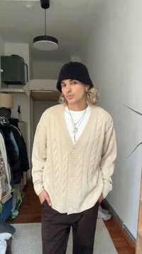 Beige kabelstrikket cardigan med V-hals, med knapper og en avslappet passform, kombinert med mørke bukser. En svart lue er på hodet.