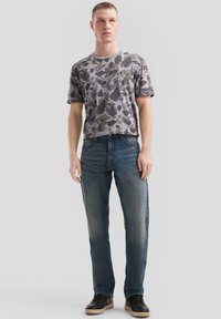 Giovane uomo in t-shirt grigia e nera mimetica, jeans blu sbiaditi e sneaker neri in piedi contro uno sfondo chiaro e uniforme.