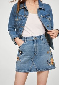 Jeansjack met twee borstzakken, gecombineerd met een blauwe jeansrok met bloemembroideries in oranje, blauw en gele accenten.