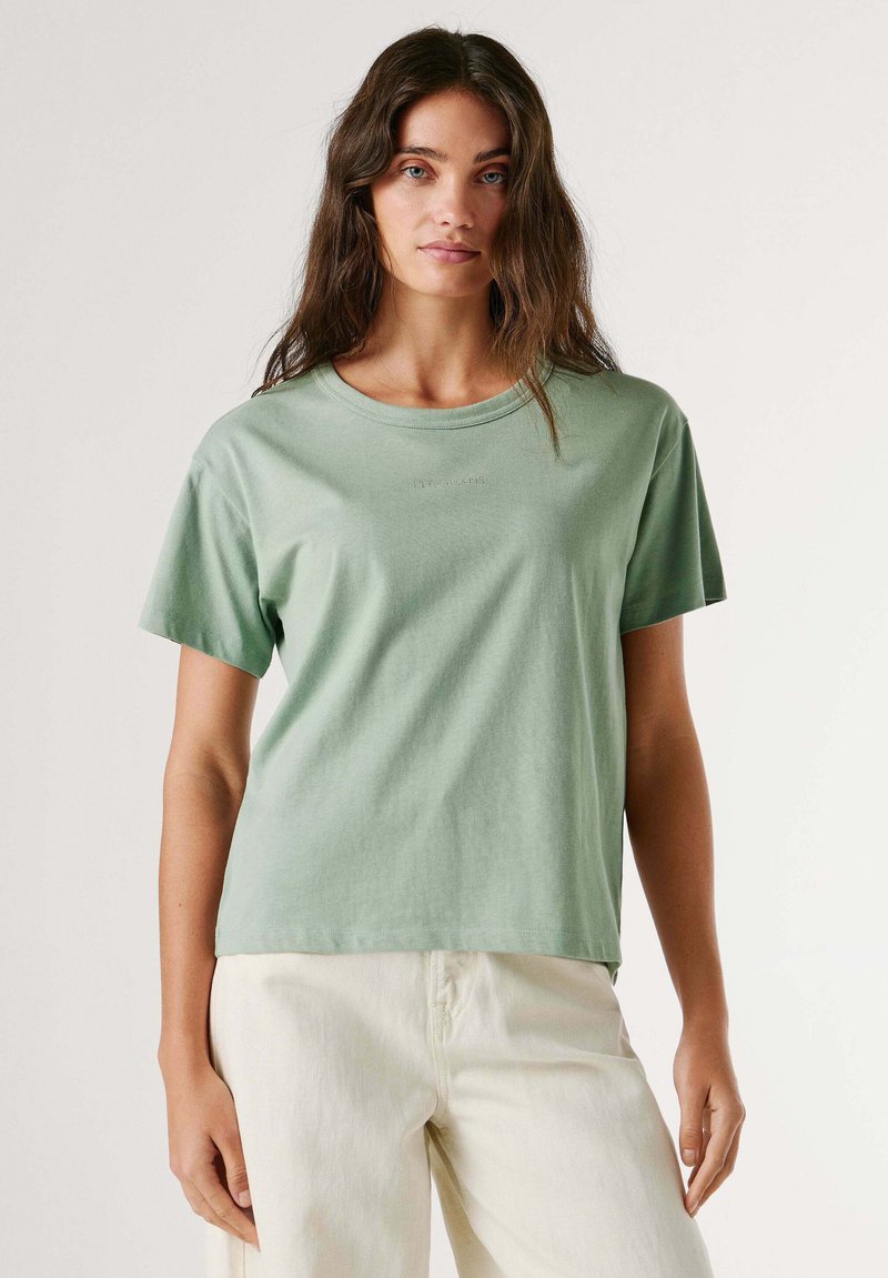 Woman with long brown hair wearing a light green short-sleeve t-shirt and off-white pants, posing against a plain light background.