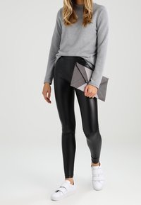 Svarta, glänsande leggings i faux läder, kombinerat med en ljusgrå långärmad tröja. En triangulär grå clutch och vita sneakers fulländar looken.
